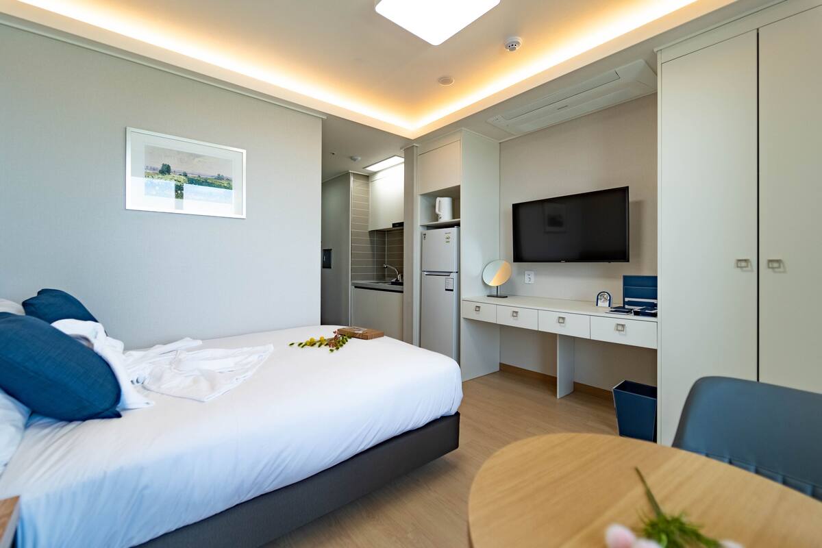 Habitación de hotel moderna con una cama grande, un escritorio con computadora, televisor y una pequeña cocina.