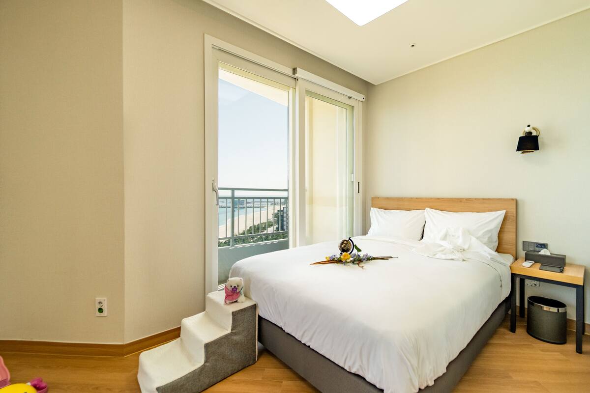 Habitación de hotel con una cama grande, una mesita de noche, un cubo de basura y una ventana con vistas al exterior.