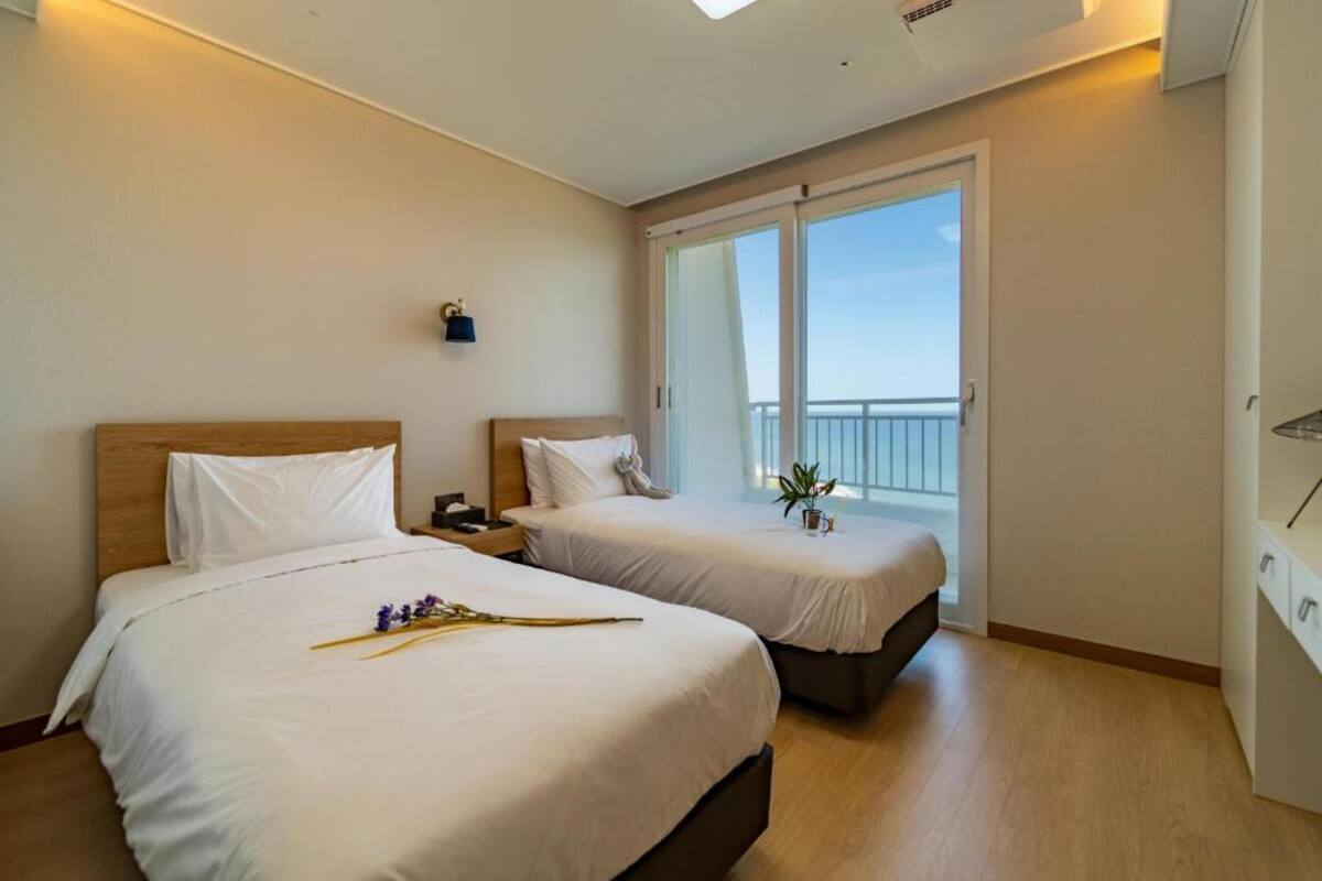 Habitación de hotel con dos camas, una mesita de noche, una pequeña mesa con una planta y un balcón con barandilla.