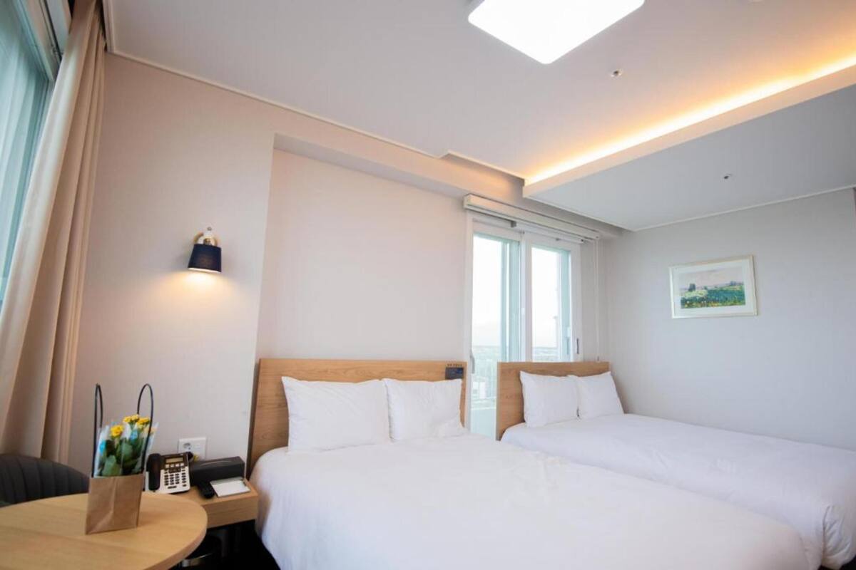 Habitación de hotel con dos camas, una mesita de noche con lámpara, una ventana y un cuadro en la pared.