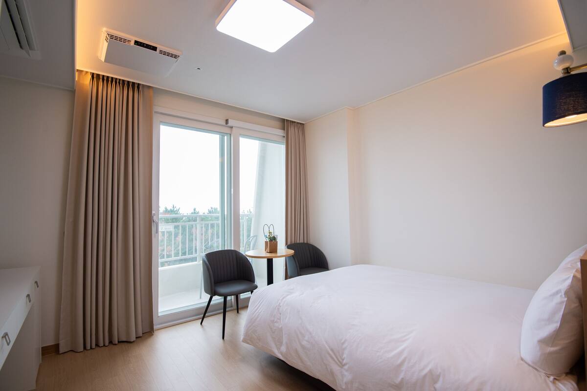 Una habitación de hotel con cama, una mesita con sillas, una ventana con cortinas y un equipo de aire acondicionado.
