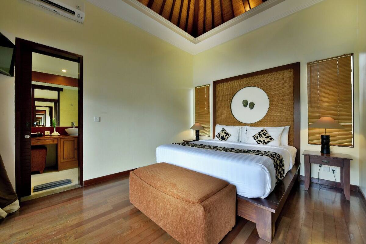 A hotel room with a large bed, a wooden headboard, a bedside table, and a bench.