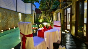 An outdoor dining area with tables set for two, covered with white tablecloths and red sashes, surrounded by stone walls and a small water feature. - The Khayangan Dreams Villa Seminyak  (Seminyak)