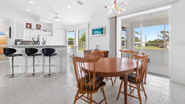 A modern kitchen with white cabinets, a dining area with a wooden table and chairs, and a large window with a view of greenery. - Cheerful & Vibrant Home Near Mineral Springs (North Port)