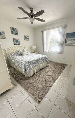 Um quarto com uma cama, um ventilador de teto, uma janela com persianas e obras de arte nas paredes.