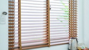 Una habitación con una ventana protegida por persianas horizontales, un escritorio blanco, una tetera y una maleta negra.