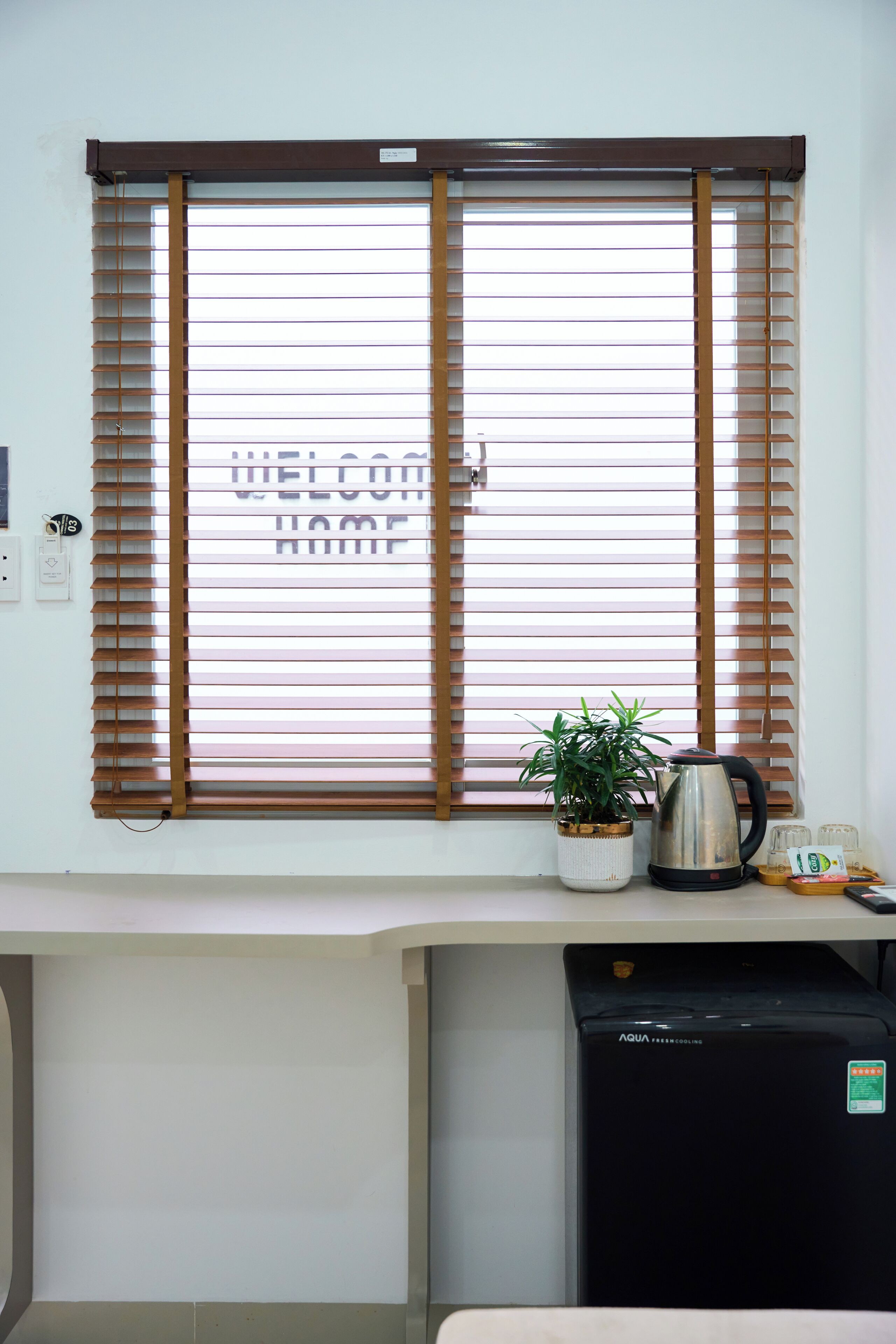 A window with horizontal blinds, a potted plant, a kettle, and a refrigerator.