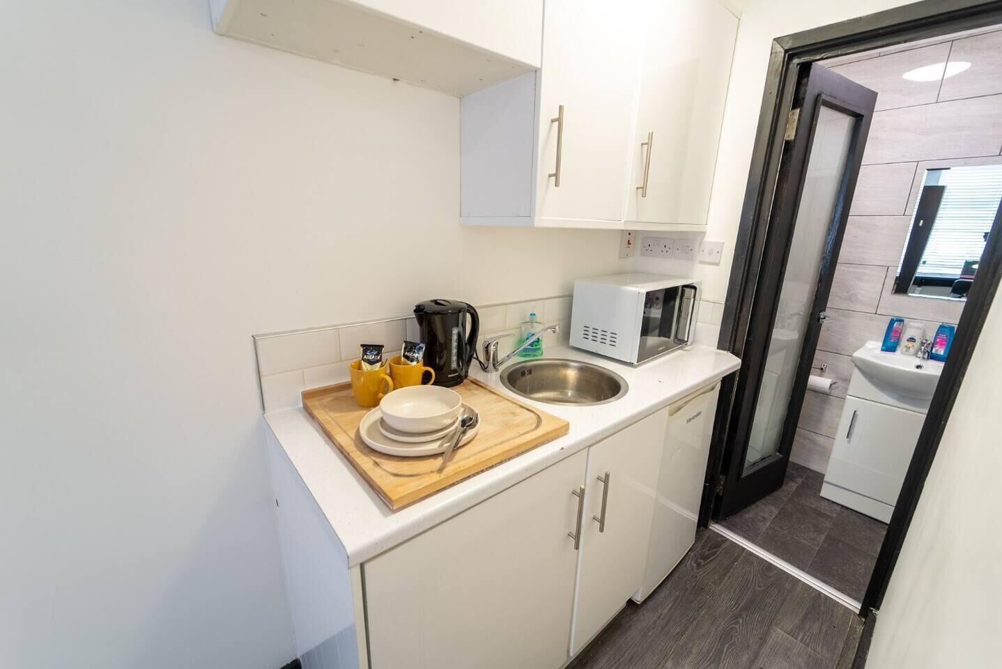 A compact kitchen with white cabinets, a microwave, a sink, and a small countertop with a cutting board, plates, and cups.