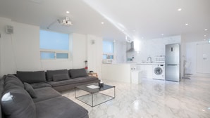 A modern living room with a grey sectional sofa, a glass coffee table, and a kitchen area with a washing machine and refrigerator. - The Wave Pension (Busan)