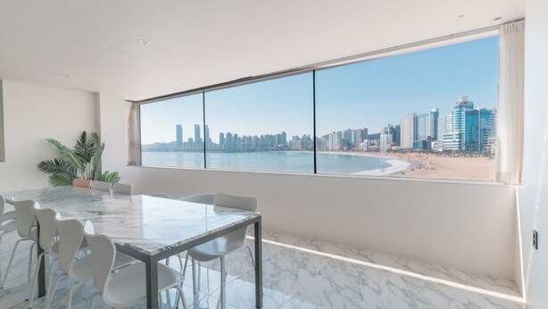 A modern room with a large window overlooking a beach and city skyline. - The Wave Pension (Busan)