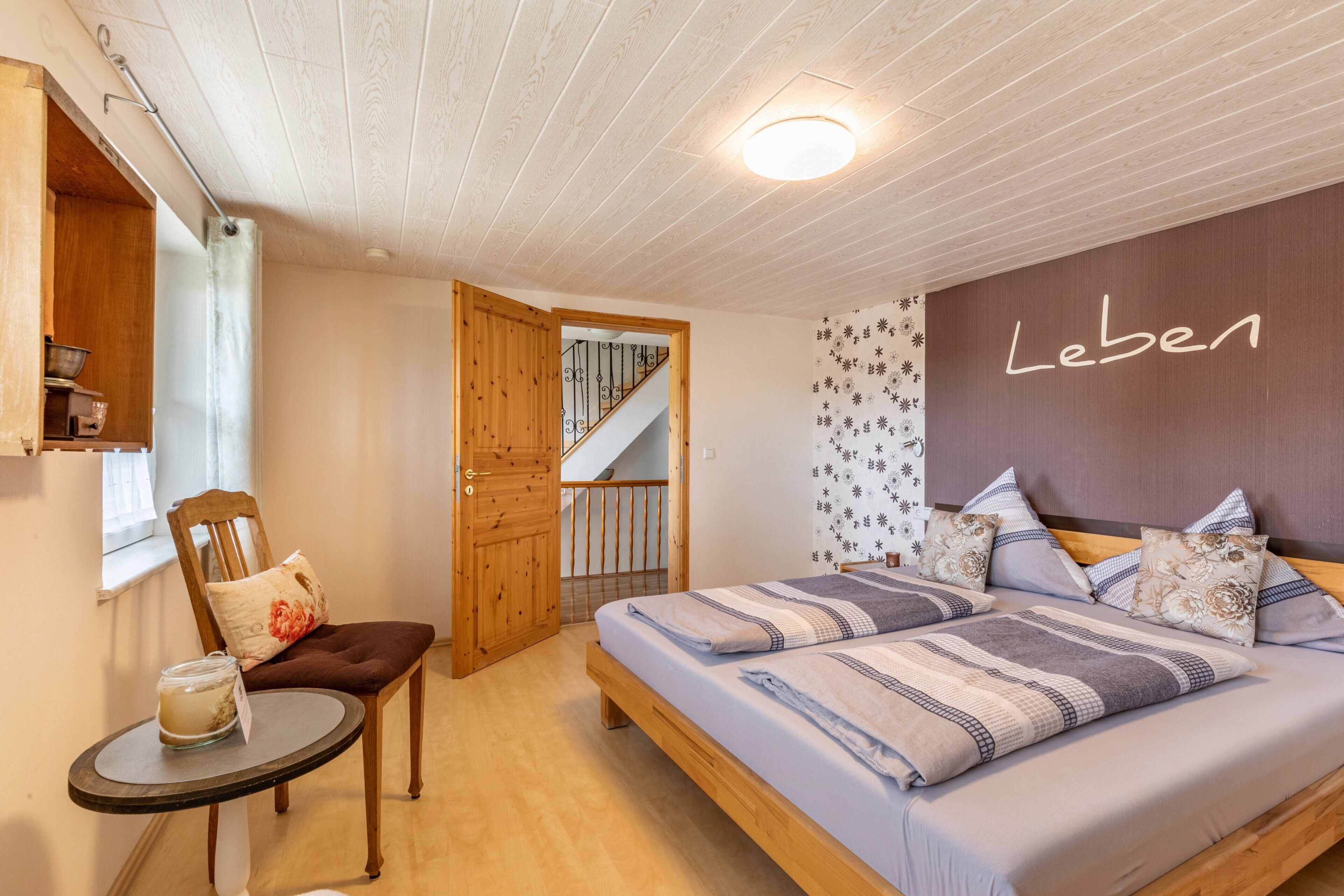A bedroom with a bed, a chair, a small table, a wooden door, a staircase, and a wall with the word 'Leben' written on it.