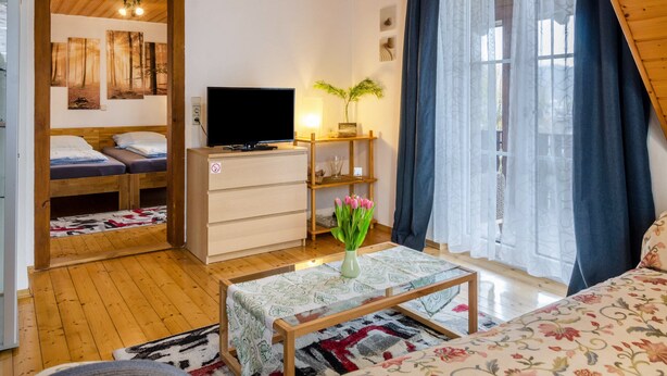 Una habitación acogedora con una cama, un televisor, un aparador de madera, una mesa con un jarrón de flores y una ventana con cortinas.