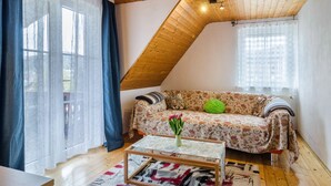 Una habitación acogedora con techo inclinado, un sofá floral, una mesa de centro con un jarrón y una alfombra estampada.