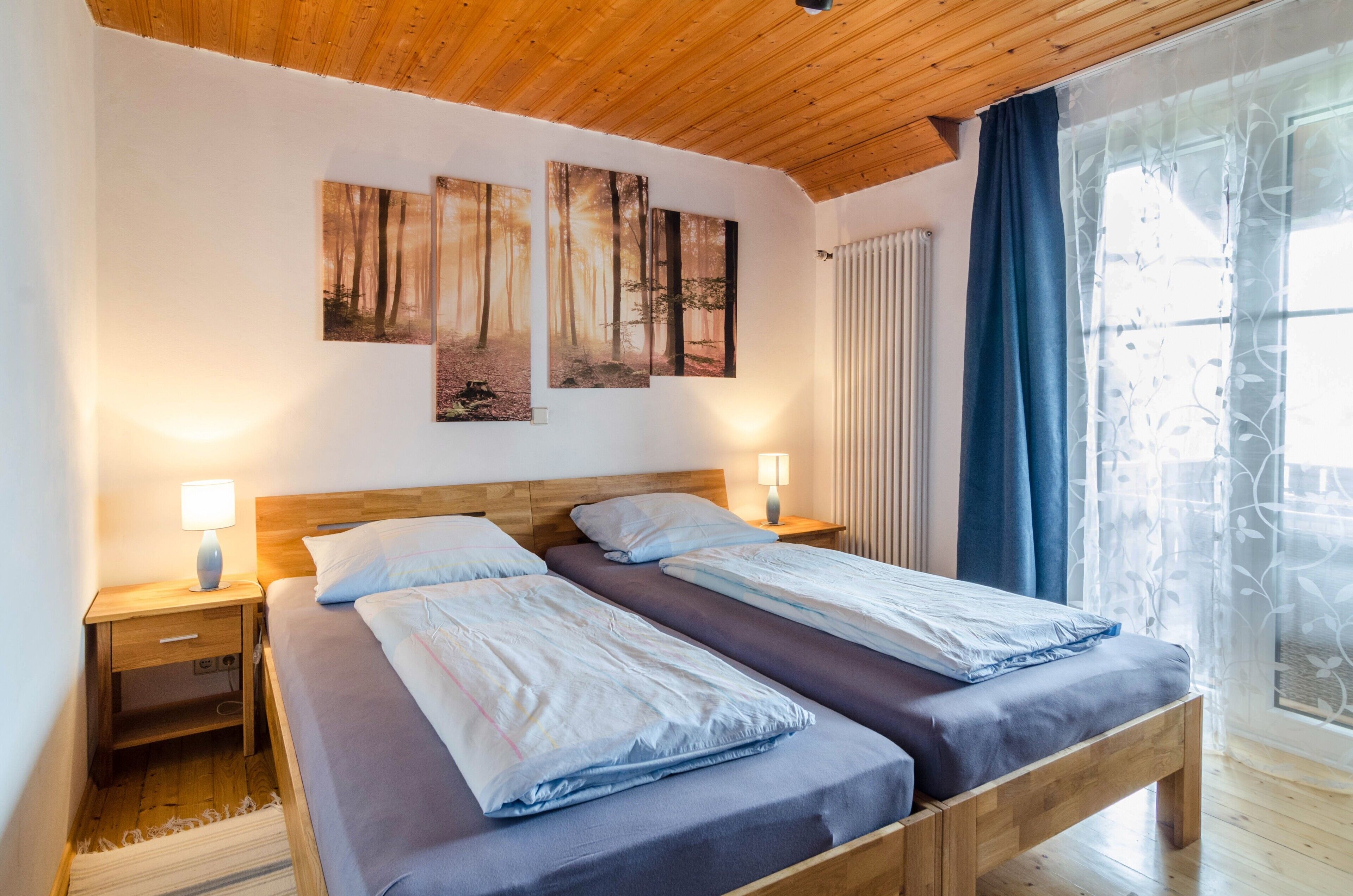 Ein Doppelbett mit hölzernem Kopfteil, zwei Nachttische mit Lampen sowie ein Fenster mit Vorhängen.