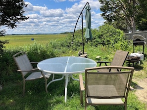 A round glass table with two wicker chairs, a green umbrella, and a barbecue grill in a grassy outdoor area with a view of a field and trees. - One-Bedroom Apt in Hampton NH! (Hampton)