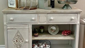 Un aparador blanco de estilo vintage con un espejo decorativo, un letrero de cafetería, una cafetera y un frutero escalonado.