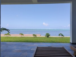 Vista desde una habitación con un banco de madera y suelo de piedra, que da a un área verde con árboles pequeños y un mar azul cristalino.