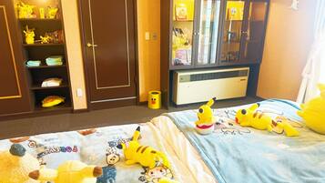 A hotel room with a bed featuring a Pikachu-themed bedspread, a wardrobe with shelves containing various items, and a small trash can.