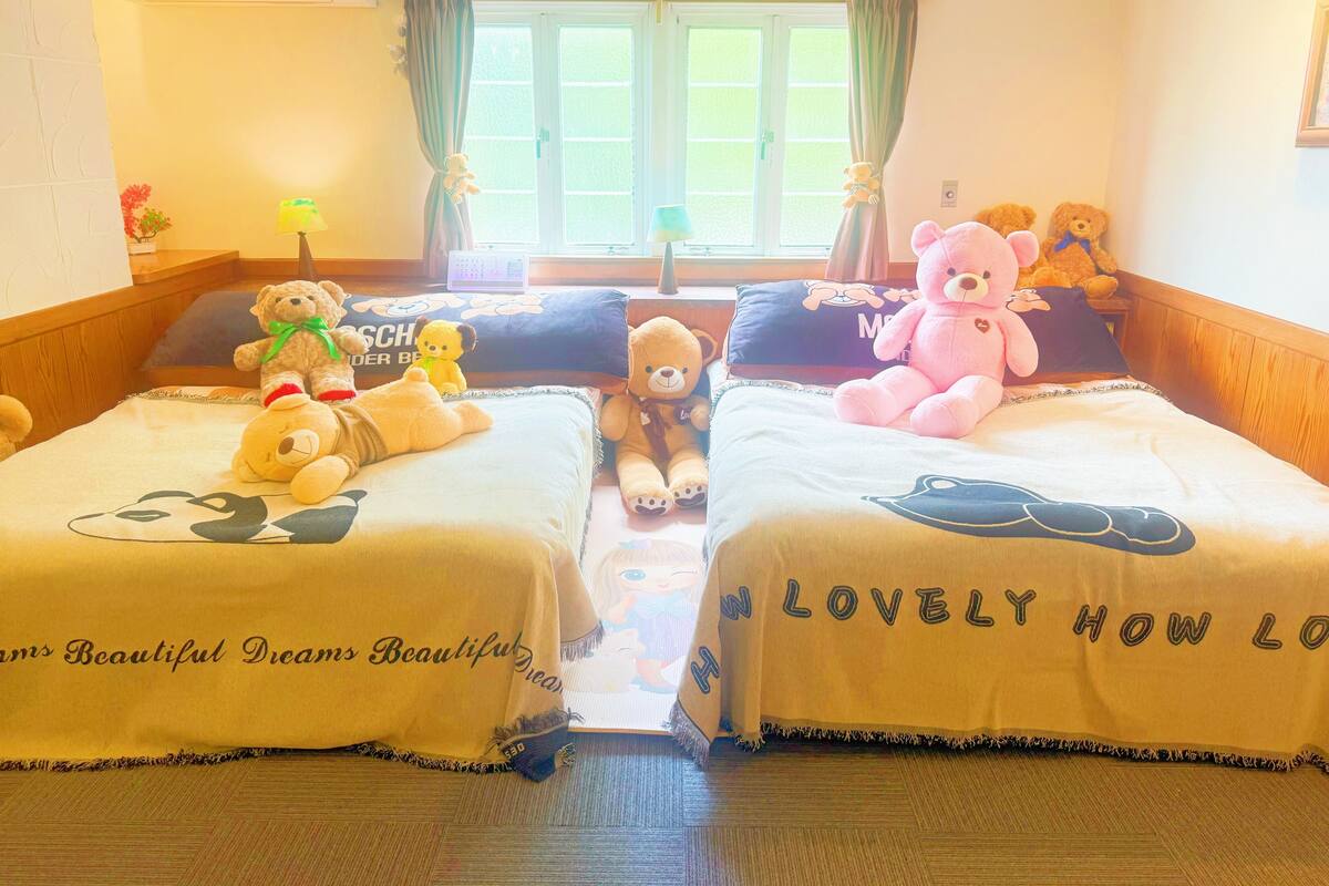 A hotel room with two beds, each with a bear-themed bedspread and pillows. There is a window with curtains, a bedside table with a lamp, and a wall-mounted air conditioner.