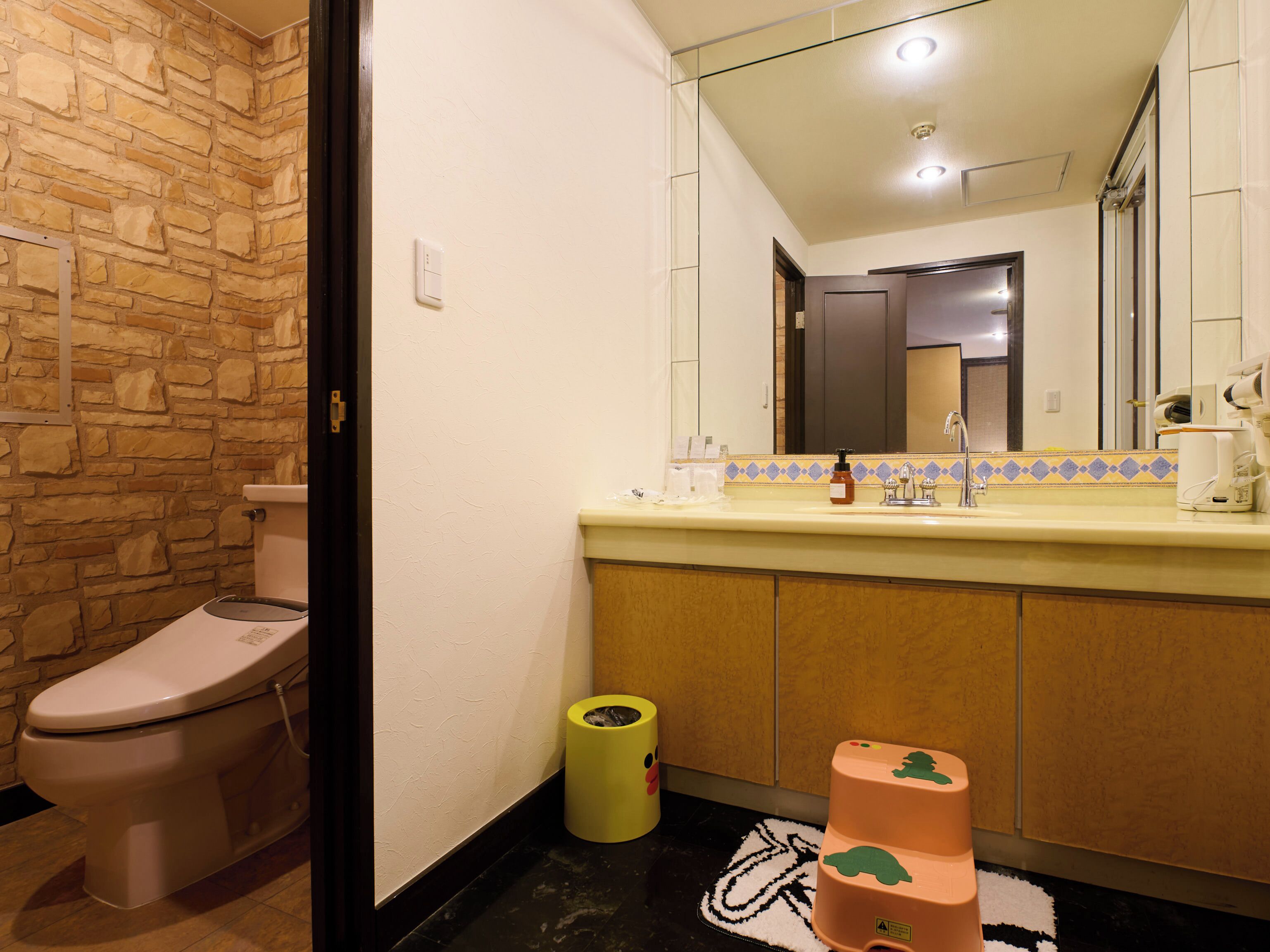 A bathroom with a stone wall, a toilet, a sink with a mirror, and a trash bin.