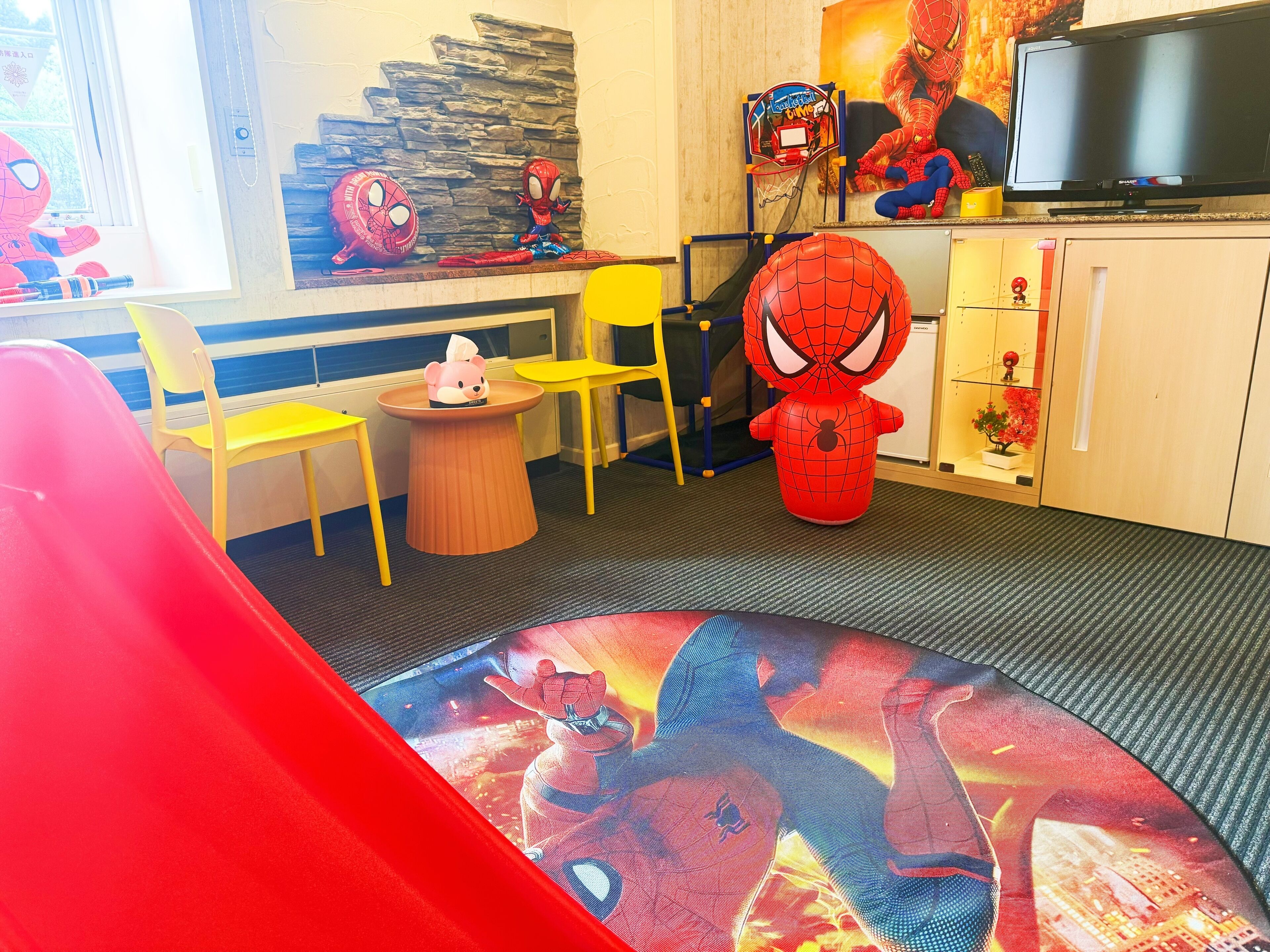 Una sala de juegos para niños con un mural de Spider-Man, un aro de baloncesto pequeño y un televisor sobre un mueble.