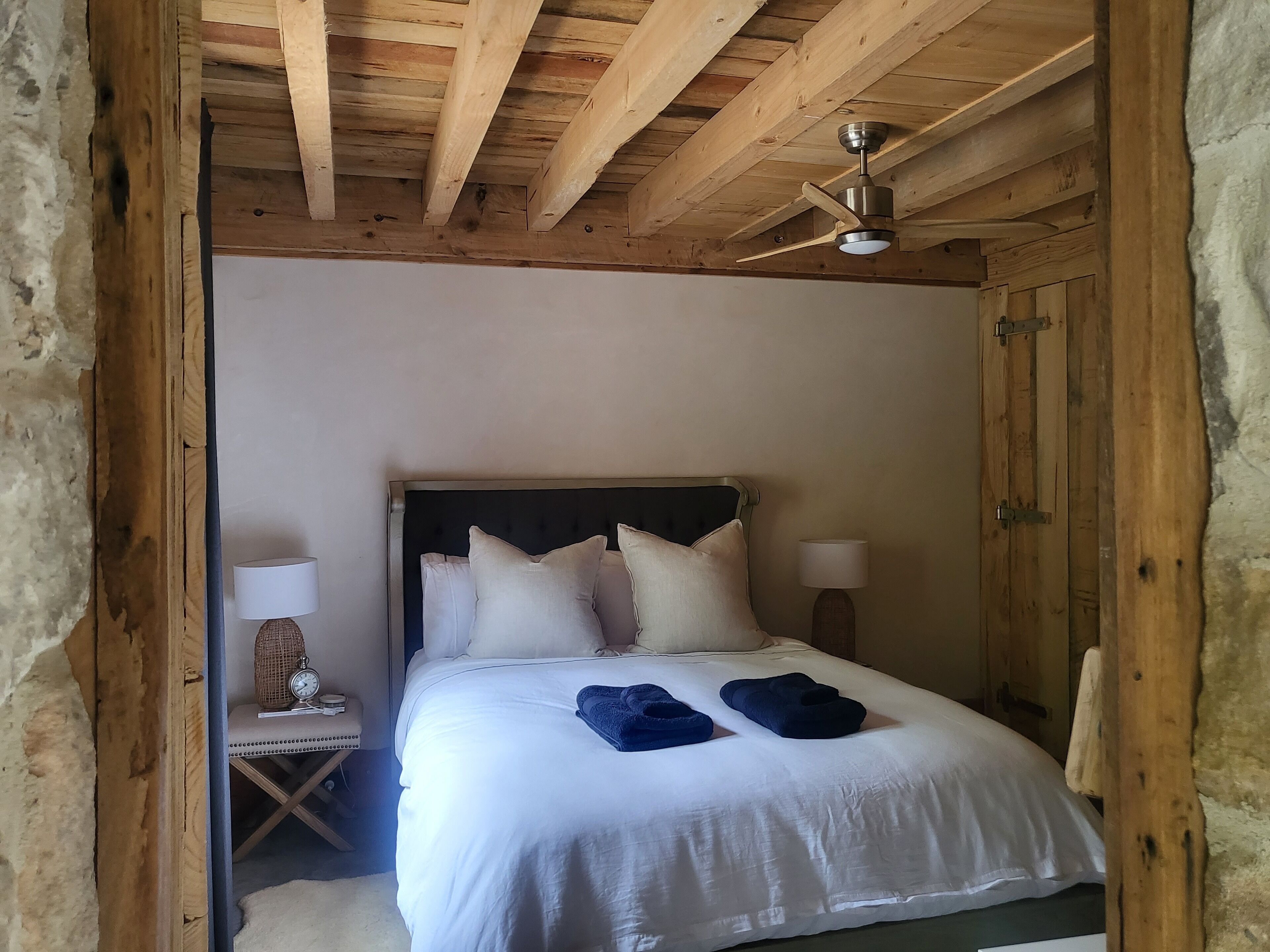 A wooden-framed bedroom with a bed, pillows, a nightstand, a lamp, and a ceiling fan.