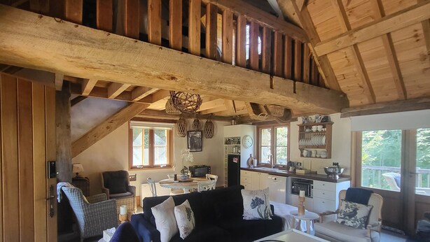 A spacious living area with a high ceiling, wooden beams, a sofa, armchairs, a coffee table, and a kitchen in the background. - Romantic Stone Cottage Overlooking Pond. 5mins to Town (Burradoo)