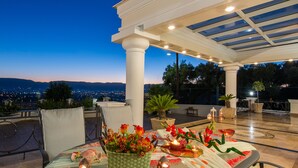 An outdoor dining area with a table set for two, a potted plant, and a view of the city at dusk.