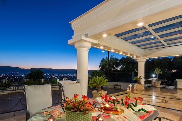 An outdoor dining area with a table set for two, a potted plant, and a view of the city at dusk.