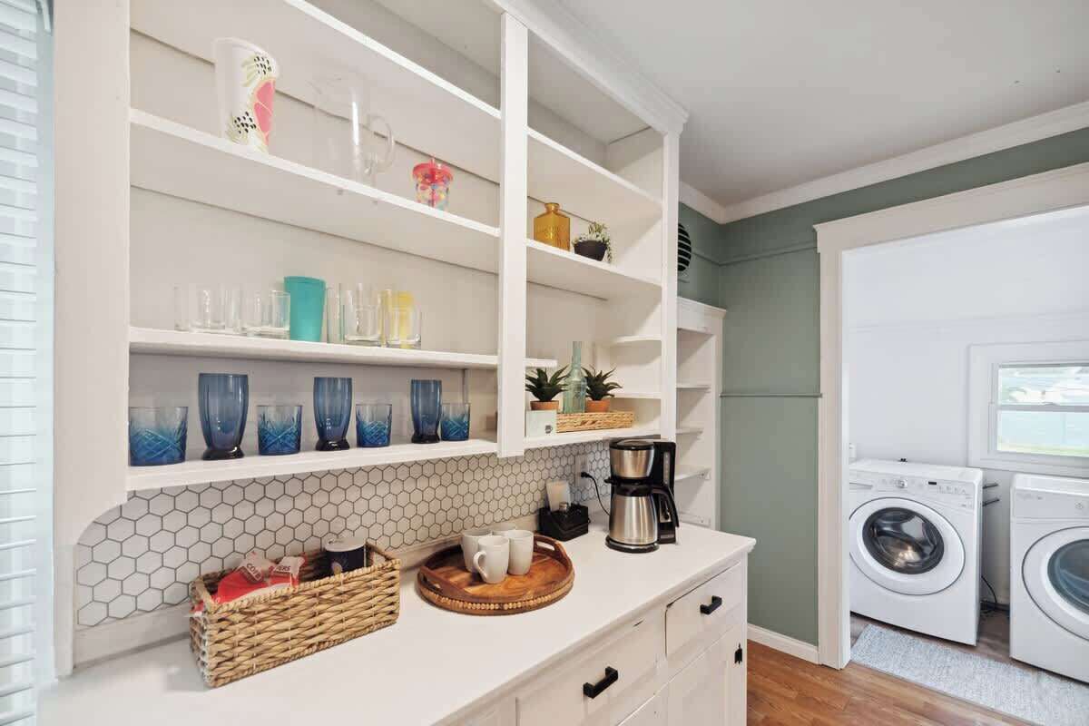 Een wasruimte met een wasmachine en droger, een koffiezetapparaat op het aanrecht, en planken met diverse spullen, waaronder drinkglazen en manden.