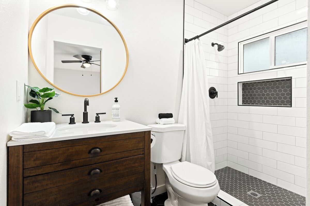 A bathroom with a white vanity, a round mirror, a ceiling fan, a toilet, and a shower with a glass door.