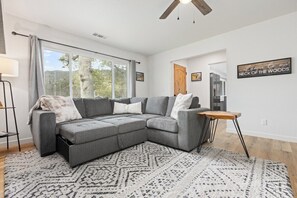 A modern living room with a sectional sofa, a coffee table, and a rug. There is a window with curtains, a ceiling fan, and a wall-mounted sign. - The Big Dipper between Kaweah River & Skyline, SNP (Three Rivers)