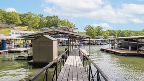 Een houten steiger die leidt naar een overdekte boothelling met een klein tuinhuisje, omgeven door kalm water en aangemeerde boten, met huizen en groen in de achtergrond.