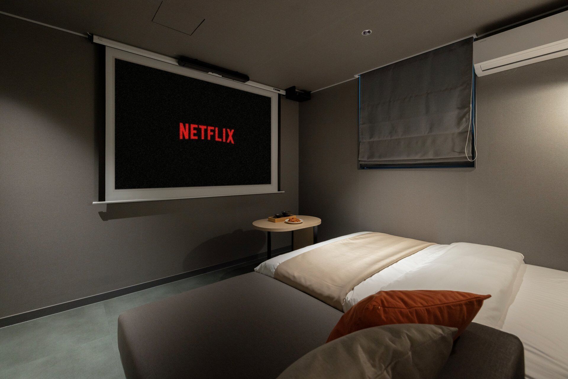 Een kamer met een bed, een klein rond tafeltje, een aan de muur bevestigde televisie waarop 'NETFLIX' staat, en een raam met een luxaflex.