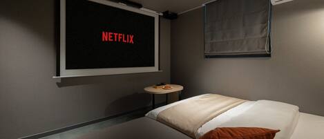 Een kamer met een bed, een klein rond tafeltje, een aan de muur bevestigde televisie waarop 'NETFLIX' staat, en een raam met een luxaflex.