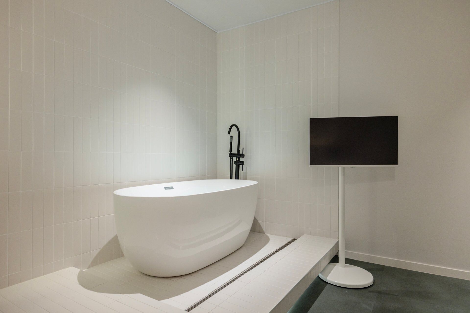 A modern bathroom with a freestanding bathtub, a black faucet, and a flat-screen TV mounted on the wall.