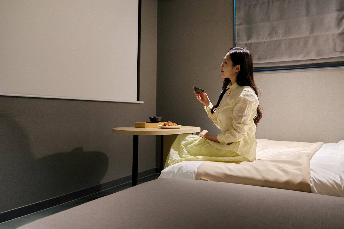 Une personne est assise sur un lit dans une pièce où se trouvent un écran de projection, une petite table avec une assiette de nourriture et une fenêtre avec des stores.