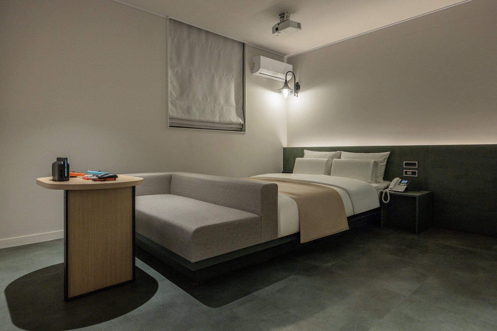 A modern hotel room with a bed, a small bedside table, a chair, a desk with a lamp, and an air conditioning unit.