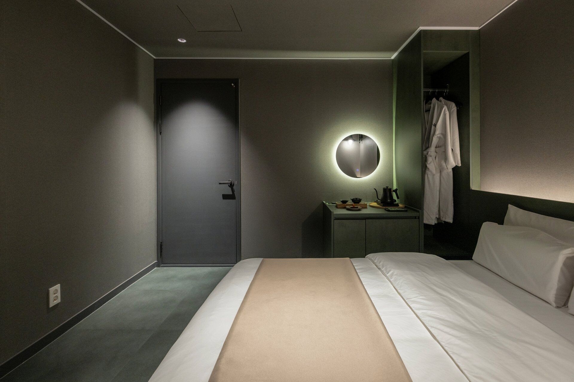 Une chambre d’hôtel moderne, équipée d’un lit, d’une table de chevet avec une lampe, d’un miroir et d’un peignoir suspendu à un porte-manteau.