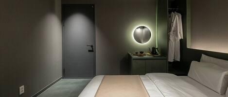 Une chambre d’hôtel moderne, équipée d’un lit, d’une table de chevet avec une lampe, d’un miroir et d’un peignoir suspendu à un porte-manteau.