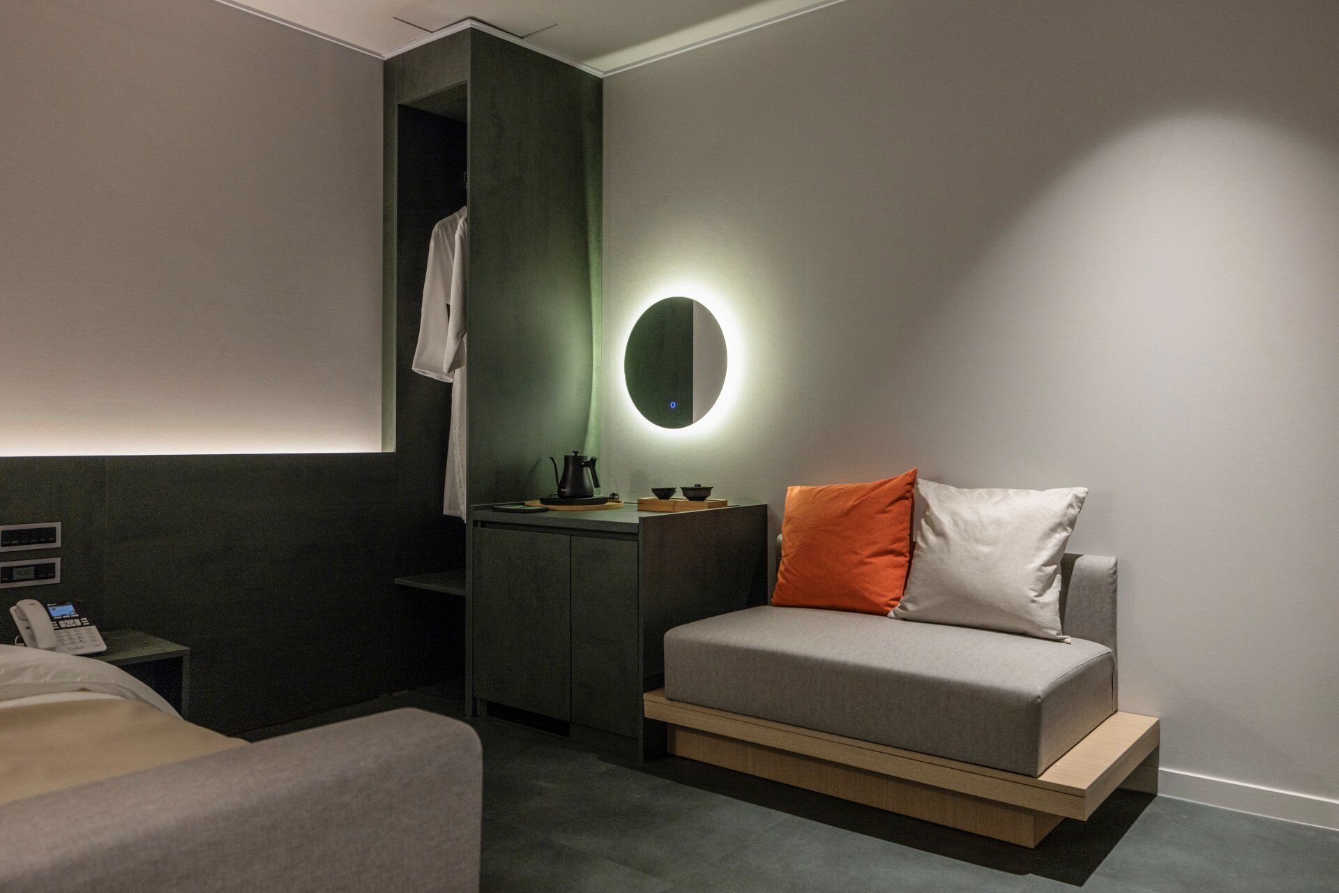 A modern hotel room with a bed, a bedside table with a phone, a small desk with a lamp, and a wall-mounted round mirror.