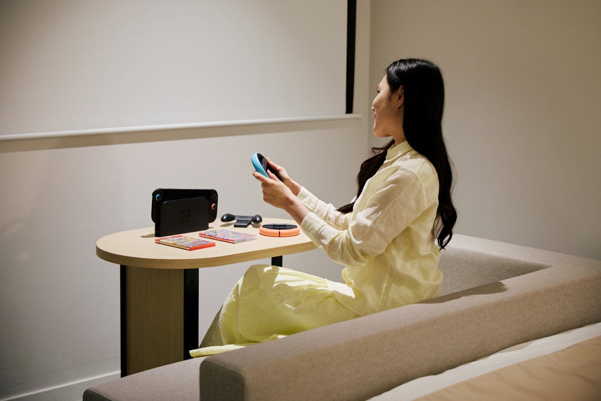 A person sitting on a couch in a room with a table that has a tablet, a game controller, and some books.