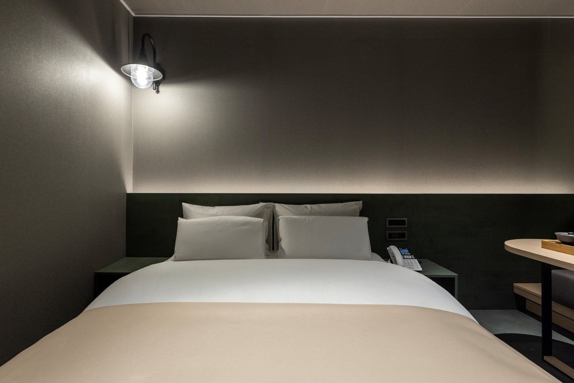 A hotel room with a bed, bedside tables, a small desk with a chair, and a wall-mounted light fixture.