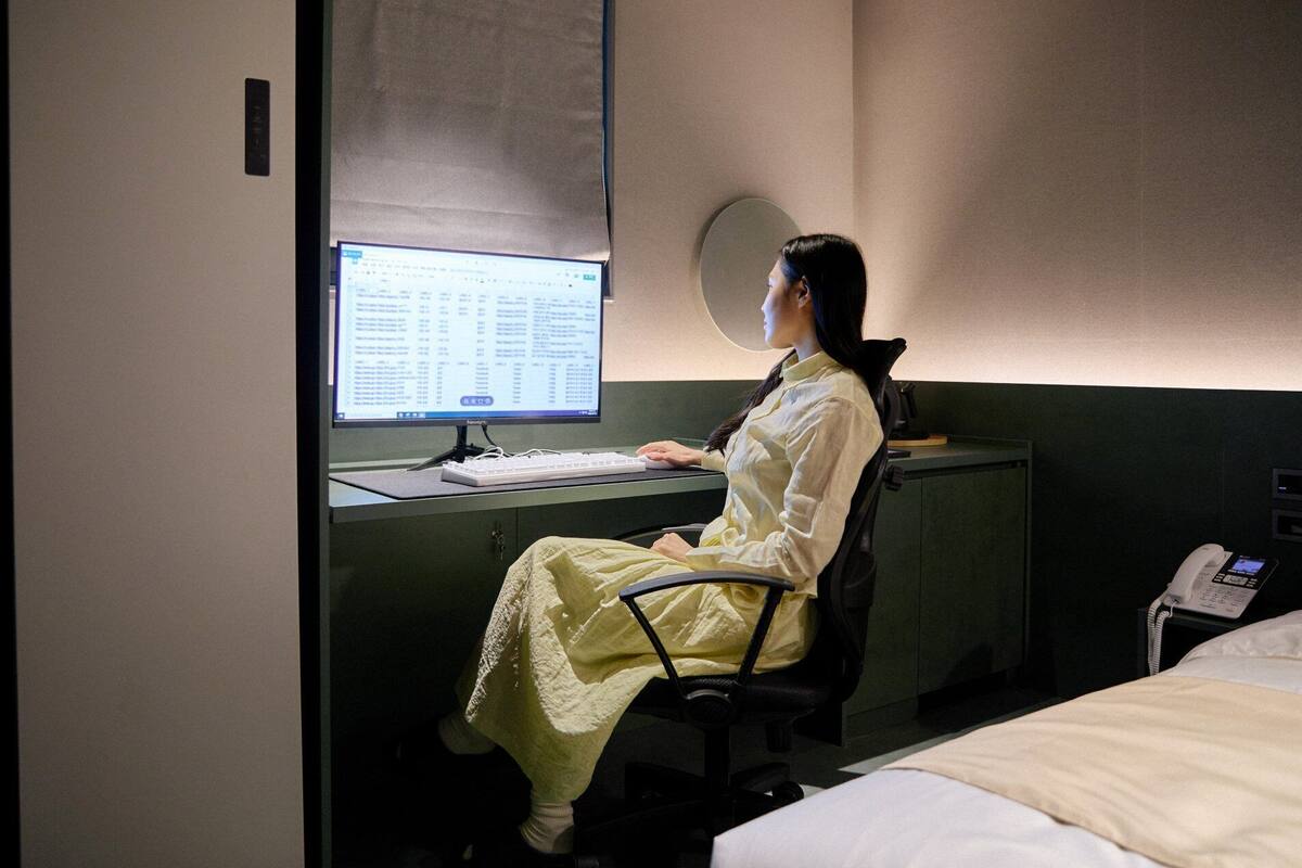Une personne assise à un bureau, avec un ordinateur, un téléphone et un lit en arrière-plan.