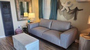 A living room with a grey sofa, a matching ottoman, a small table with a lamp, a mirror, and a decorative starfish wall hanging. - Lil Beachy (Memphis)