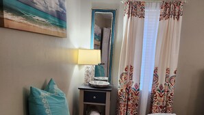 A bedroom with a bed, a nightstand, a lamp, a mirror, and a window with patterned curtains. - Lil Beachy (Memphis)