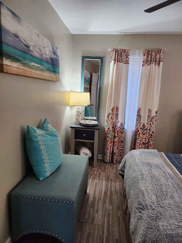 A bedroom with a bed, a nightstand, a lamp, a mirror, and a window with patterned curtains. - Lil Beachy (Memphis)
