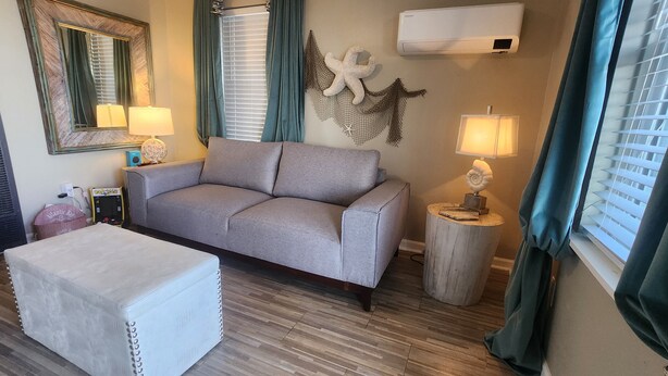 A living room with a grey sofa, a white ottoman, a round wooden side table with a lamp, a wall-mounted starfish decoration, and a large mirror. - Lil Beachy (Memphis)