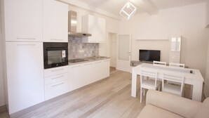 A modern kitchen with white cabinets, a black oven, and a tiled backsplash. There is a dining area with a white table and chairs, and a flat-screen TV mounted on the wall.