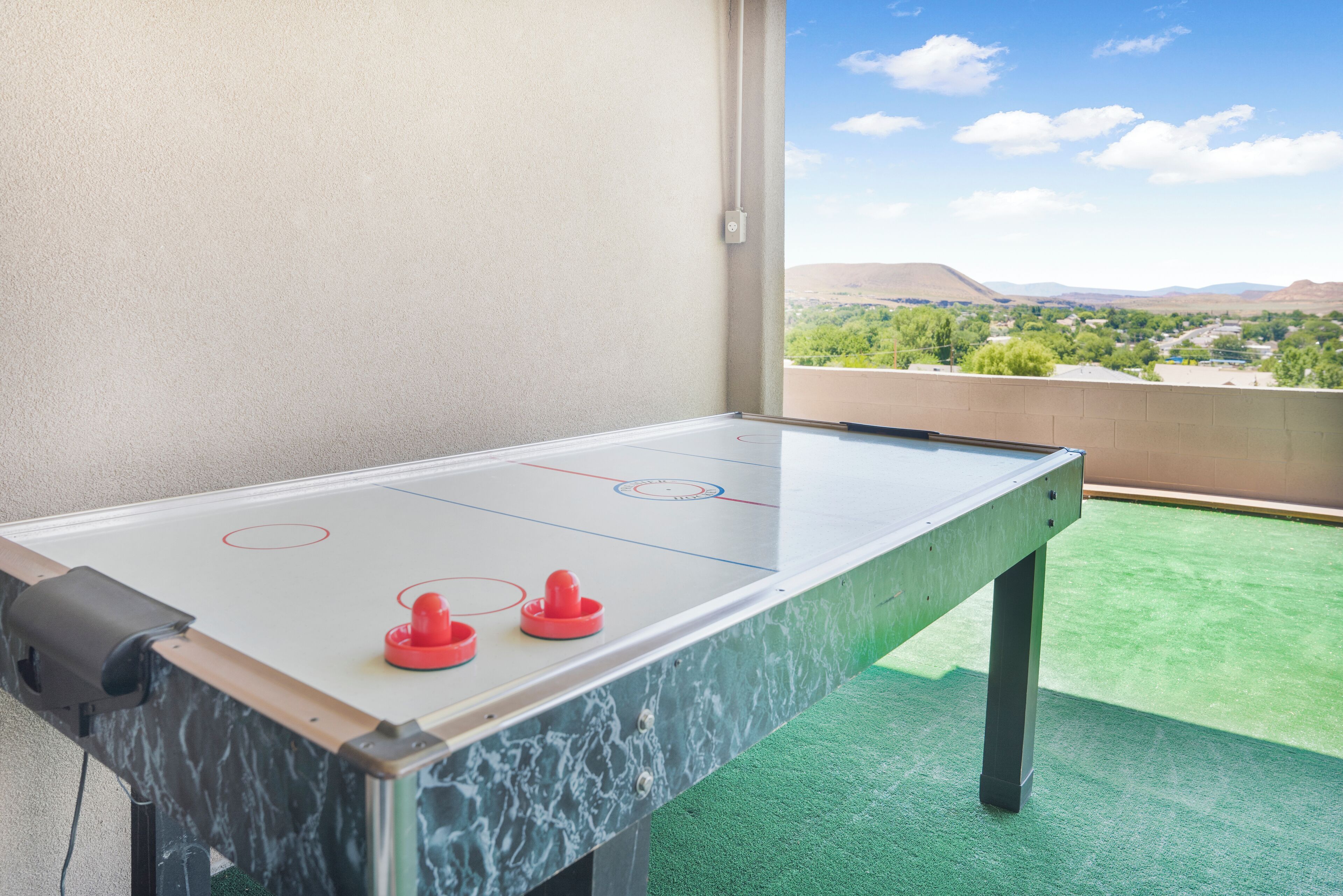Une table de baby-foot offrant une vue dégagée sur un sol recouvert d’un gazon synthétique vert, ainsi qu’un balcon avec une vue panoramique sur des collines lointaines et la verdure environnante.
